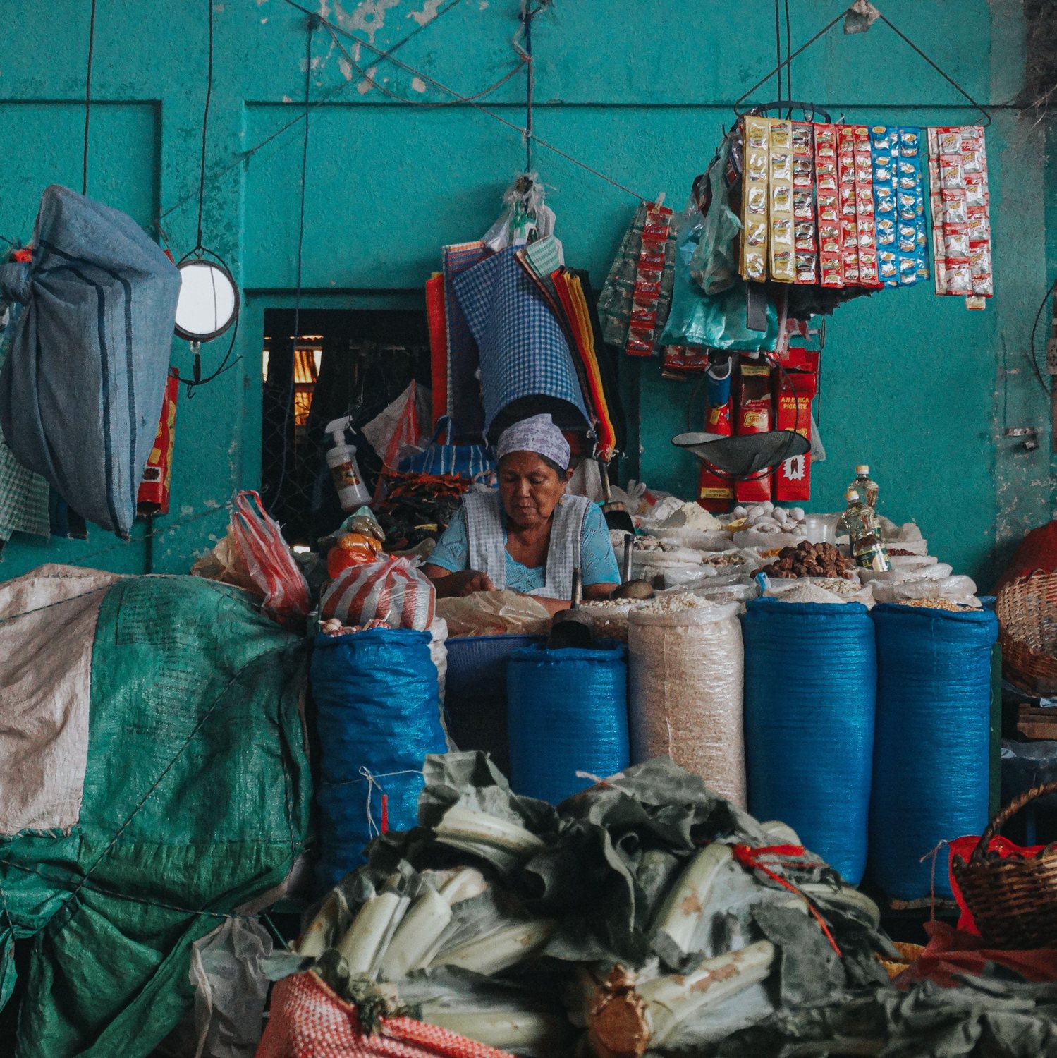 A woman tends her stall in a market hall, among blue sacks of grain against a turquoise wall