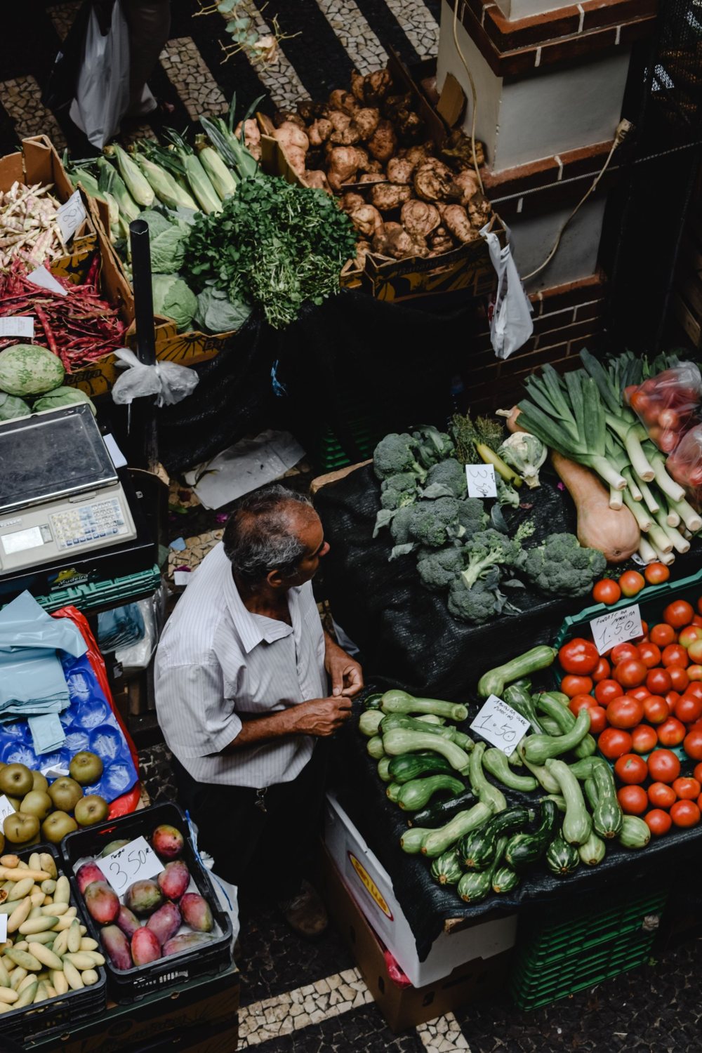 Seen from above, a man picks vegetables at a market stall full of tomatoes, broccoli and onions